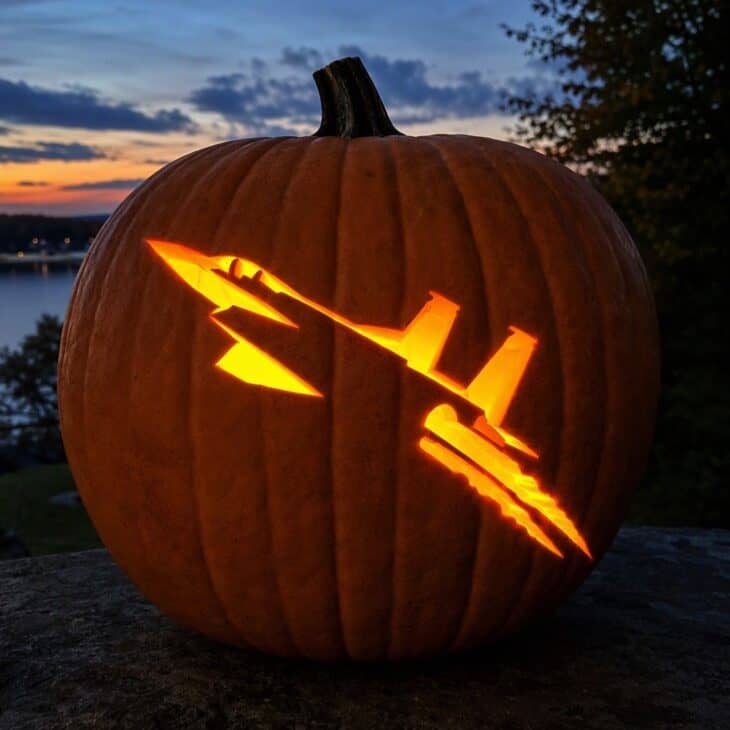 A large pumpkin carved with a glowing fighter jet silhouette diving at an angle with twin tails and an exhaust trail, outdoors at dusk.