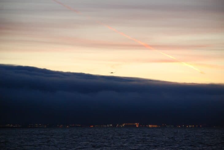 Dark fog bank over San Francisco Bay at dusk with an airliner on approach and airport lights below