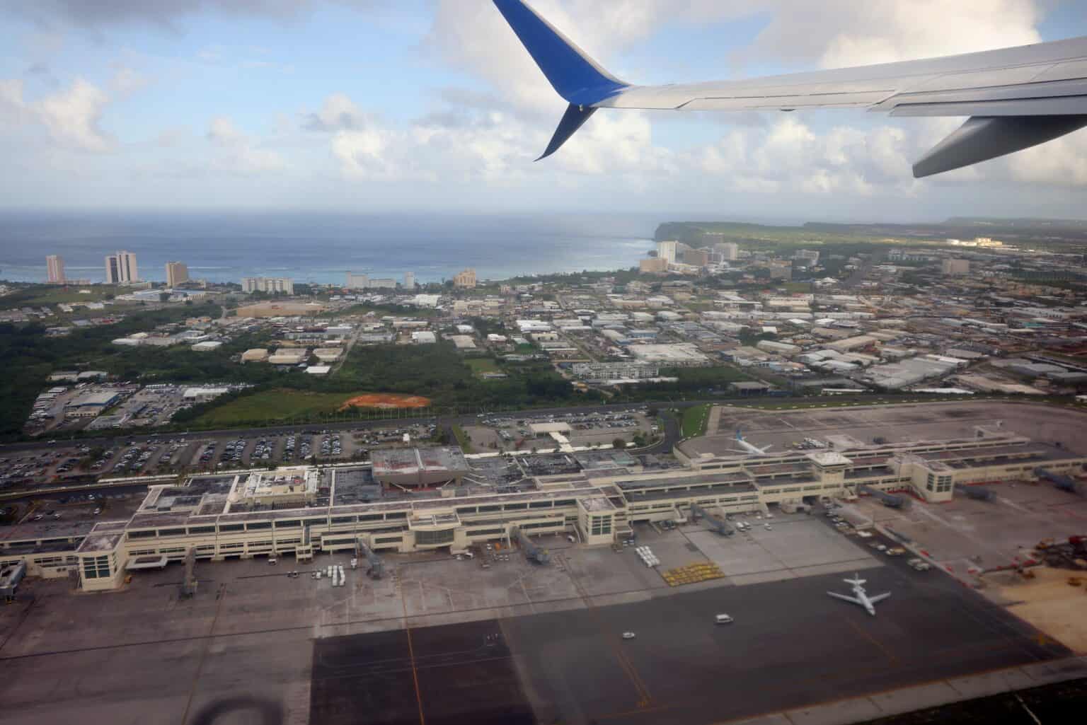 Aerial view of Guam's Antonio B. Won Pat International Airport terminal and ramp from an aircraft window, with a wing and winglet in the frame