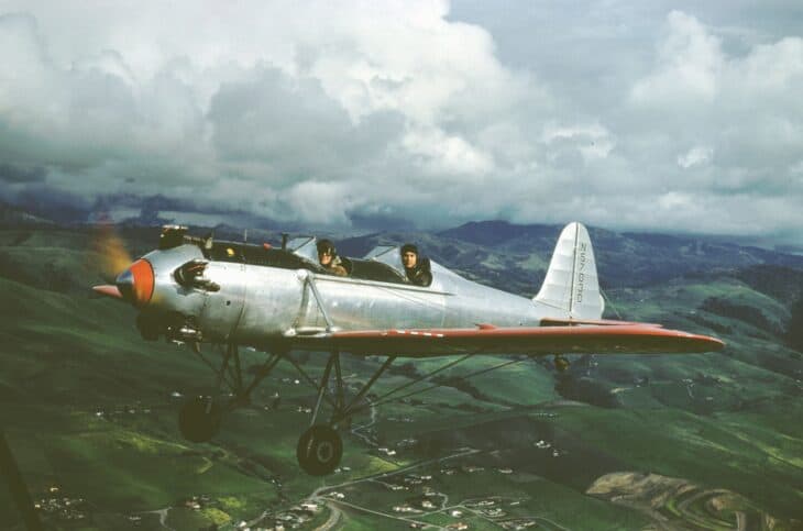 Silver Ryan PT-22 Recruit trainer N57030 with an orange nose and wing in flight over green hills under storm clouds