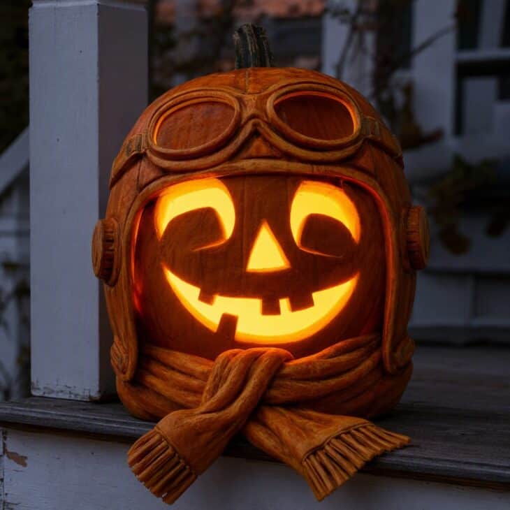 A classic jack-o'-lantern face with carved aviator cap and goggles on the forehead and a scarf around the stem.