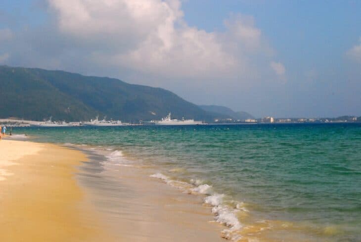 Grey warships moored across Yalong Bay beyond a sandy beach near Sanya, Hainan