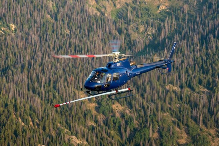 Blue Airbus H125 helicopter in flight over forested terrain