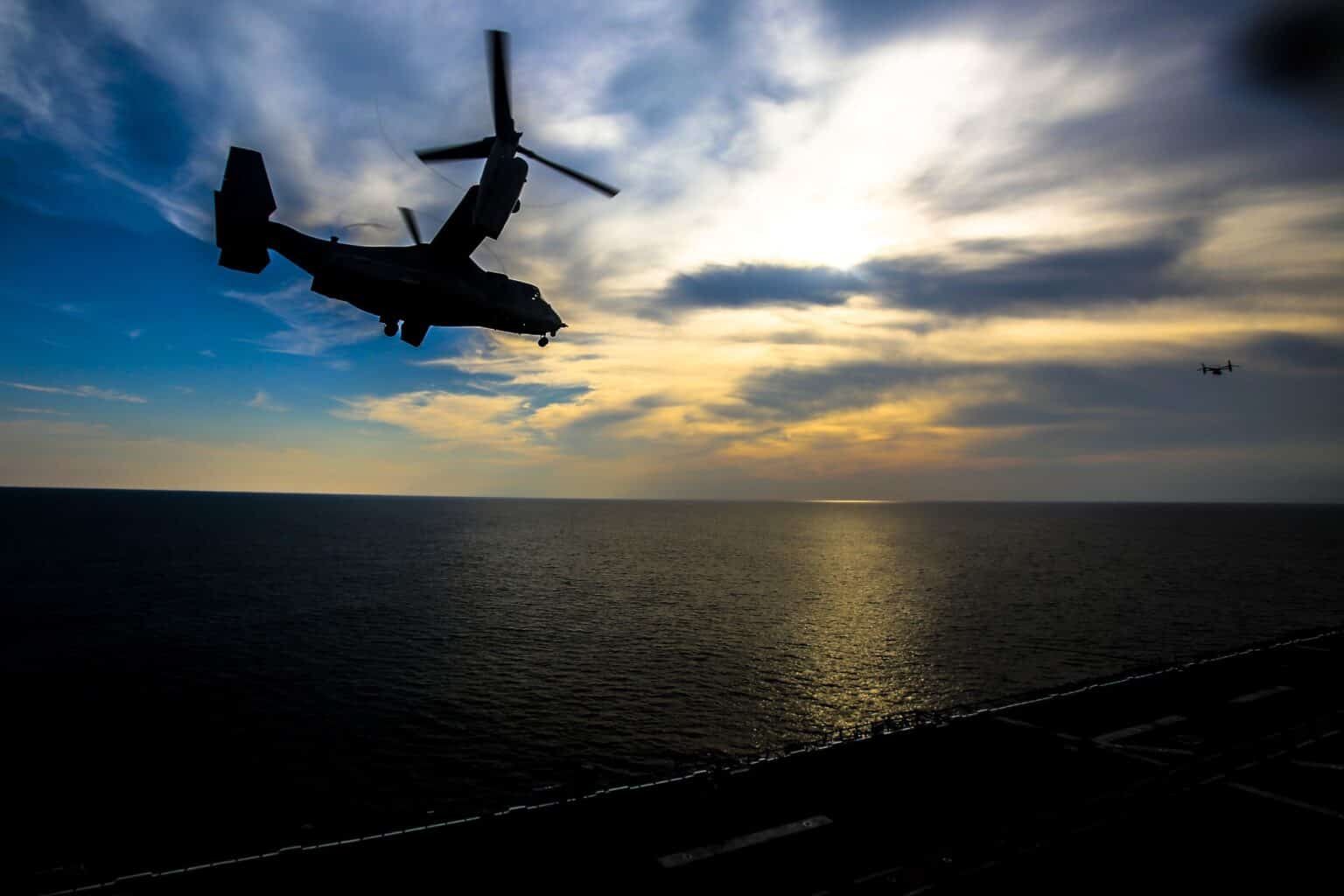 Silhouette of a Marine Corps MV-22B Osprey lifting off over the ocean against a sunset sky, with a second Osprey in the distance