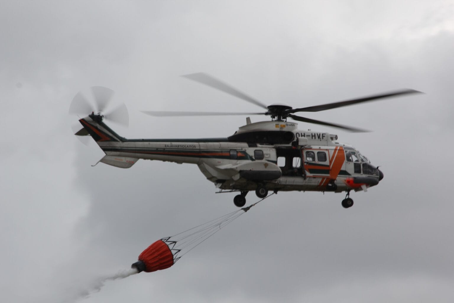 AS332 Super Puma helicopter OH-HVF flying with a red firefighting water bucket on a longline