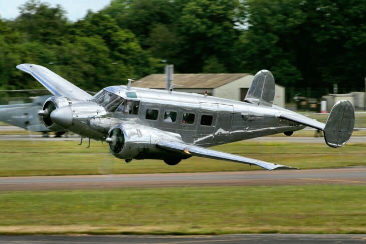 Silver Beechcraft Model 18 twin-engine airplane skimming low over a grass airfield during an air show
