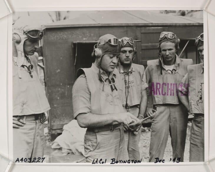 Lt. Col. Gregory Boyington in flight gear briefing Black Sheep pilots beside a hut in December 1943