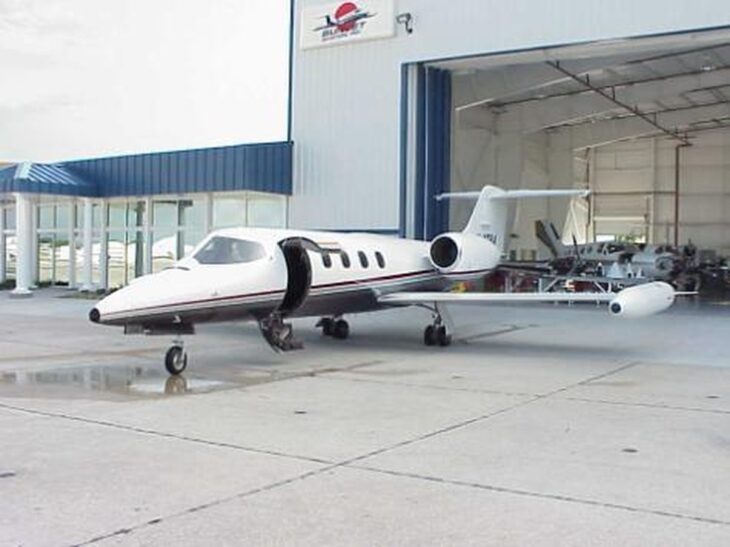 White Learjet 35 N47BA with a maroon cheatline and tip tanks parked on a wet ramp outside a Sunjet Aviation hangar, cabin door open
