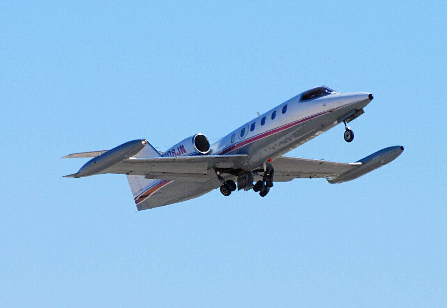 White Learjet 35A business jet with a maroon cheatline climbing in a bank against a clear blue sky, landing gear still retracting