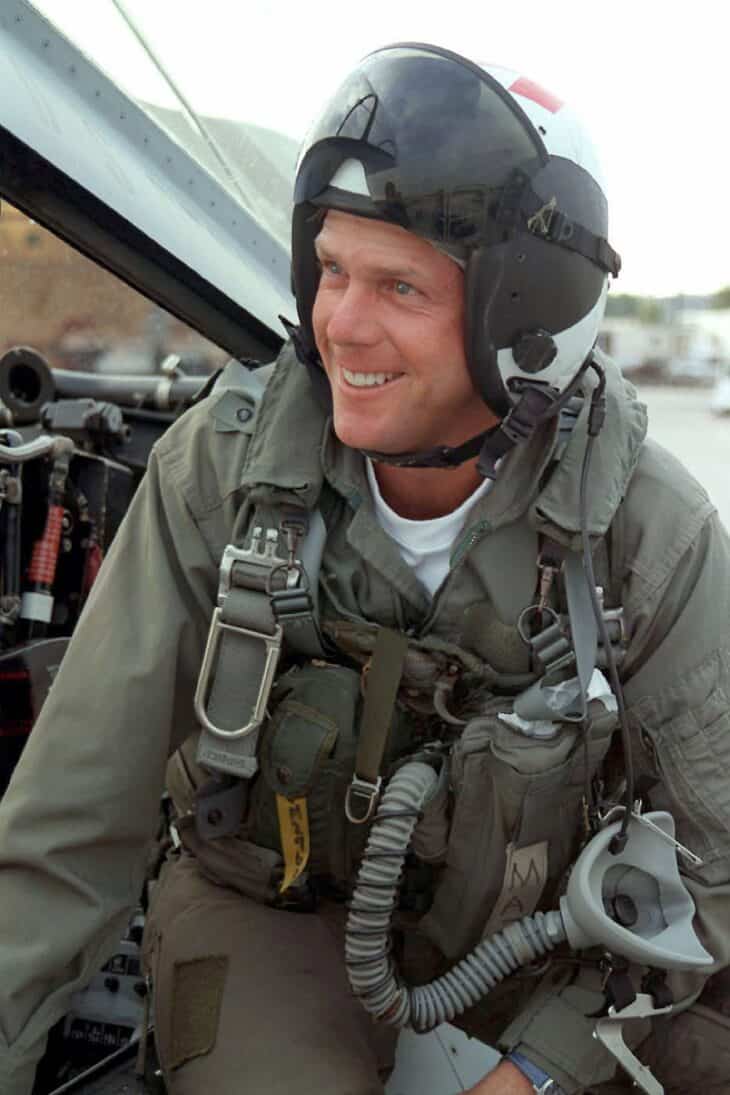 Payne Stewart smiling in a Navy flight suit, helmet and harness, an oxygen mask hanging at his chest, beside the cockpit of an F-14 Tomcat