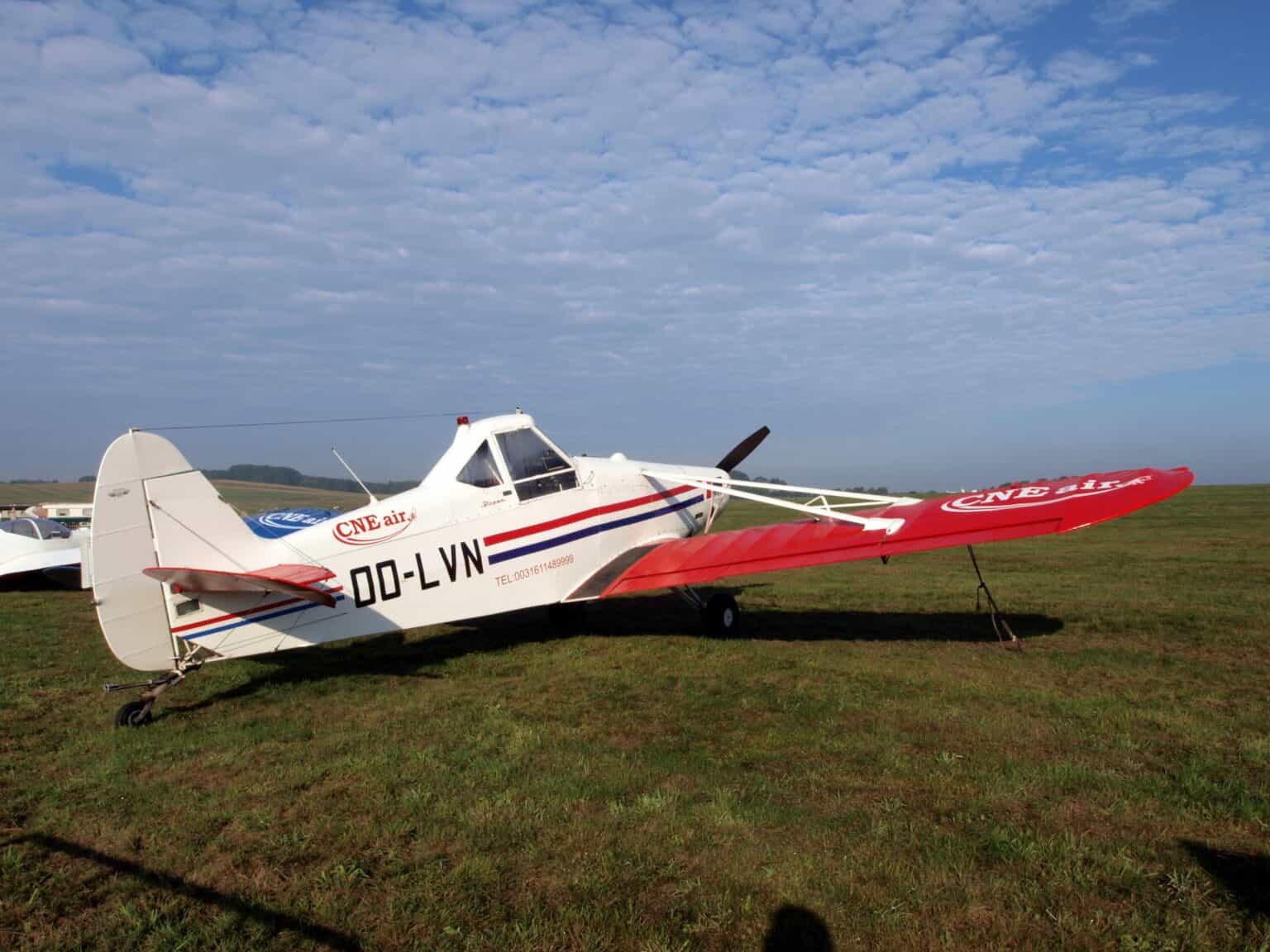 White Piper PA-25-235 Pawnee crop sprayer with a red tail parked on grass under a partly cloudy sky