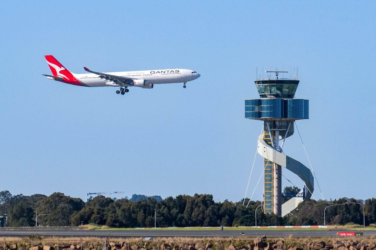 Qantas airliner with gear down flying past the Sydney Airport air traffic control tower under a clear blue sky