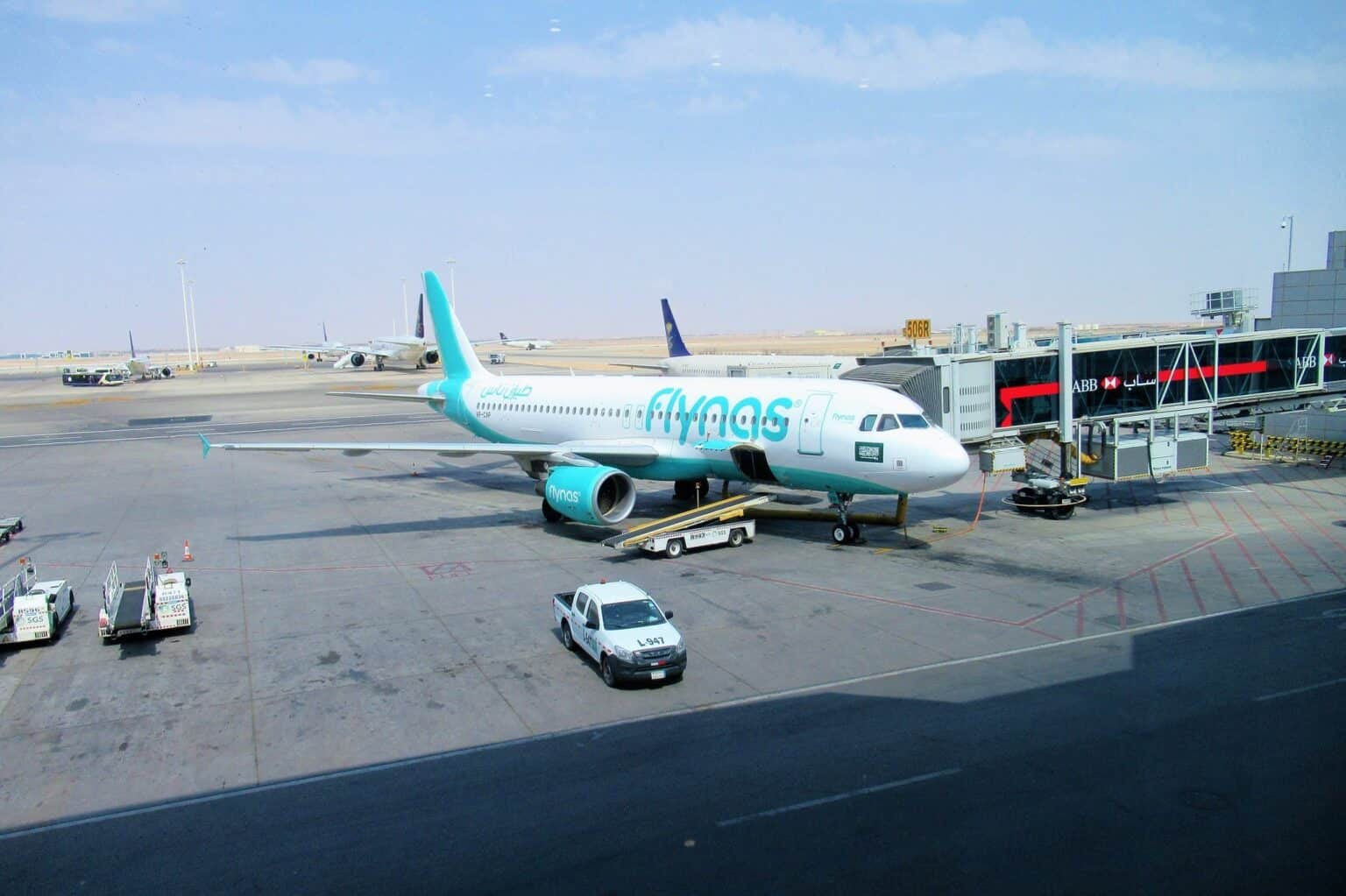 Flynas Airbus A320 on the apron at King Khalid International Airport in Riyadh with a jet bridge and Saudia tail fins behind