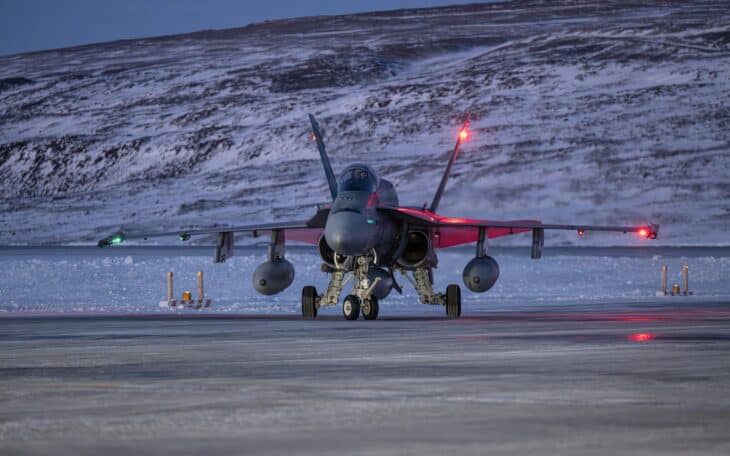 Canadian CF-188 Hornet parked on a snow-covered Arctic ramp at dusk with its afterburner lights glowing