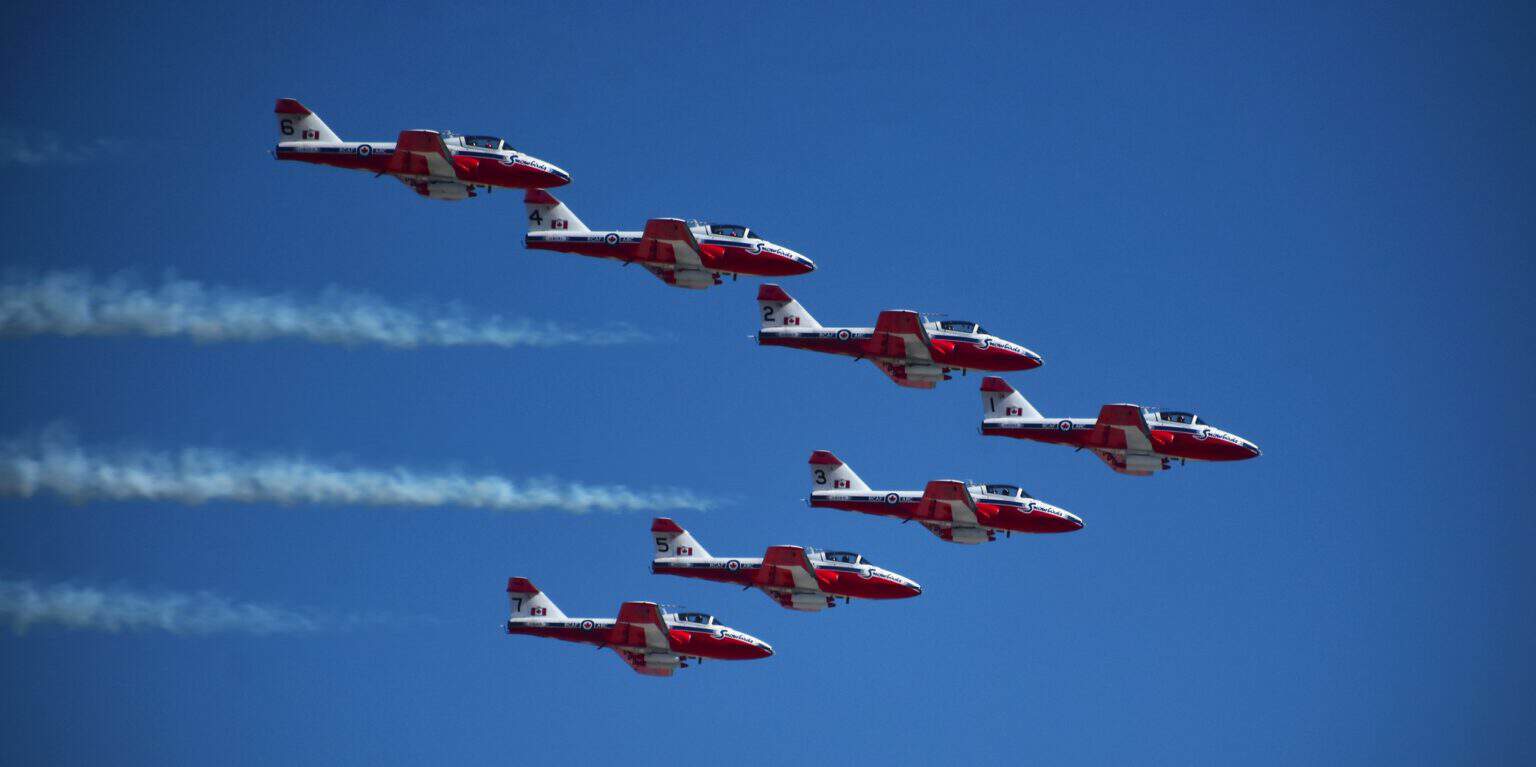Six red and white Canadair Tutor jets of the Snowbirds flying in formation trailing white smoke against a deep blue sky