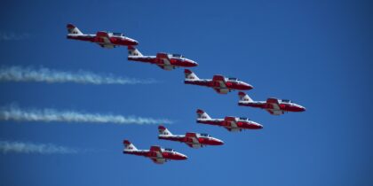 Six red and white Canadair Tutor jets of the Snowbirds flying in formation trailing white smoke against a deep blue sky