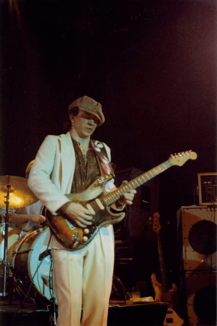 Stevie Ray Vaughan playing guitar on stage in a white suit and hat