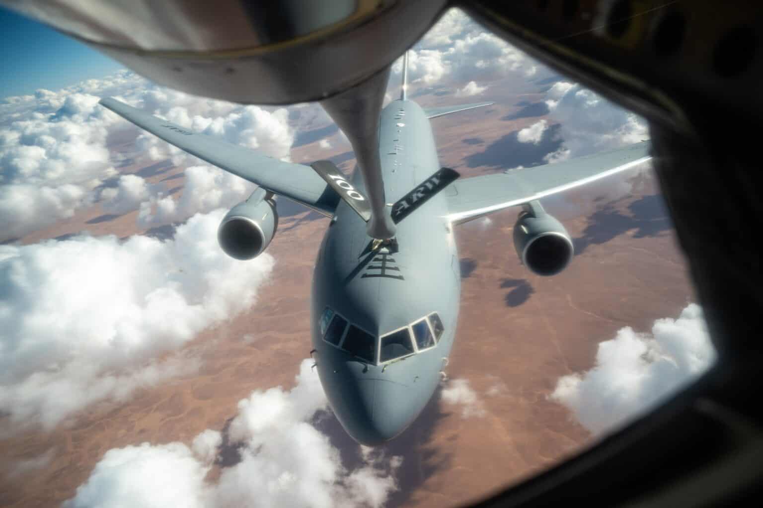 View up the refueling boom at a gray USAF KC-46A Pegasus tanker flying over desert and clouds, boom nozzle at its receptacle