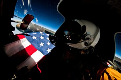 U-2 pilot in a pressure suit and helmet in the cockpit at altitude, with an American flag across the lap and the curve of the Earth beyond the canopy