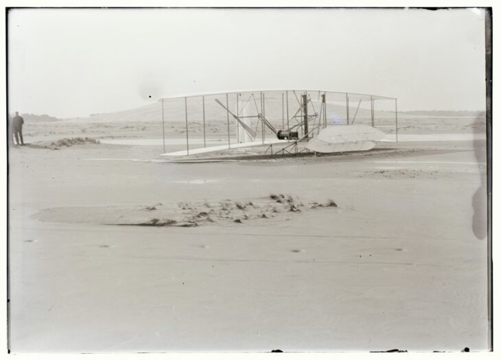The 1903 Wright Flyer resting on the sand at Kill Devil Hills after its fourth and final flight, with the dunes behind it