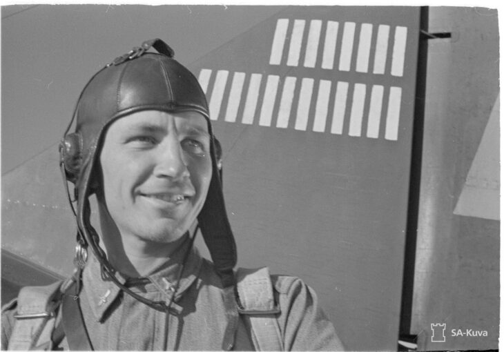 Ilmari Juutilainen in a flying helmet in front of a fighter tail marked with victory stripes